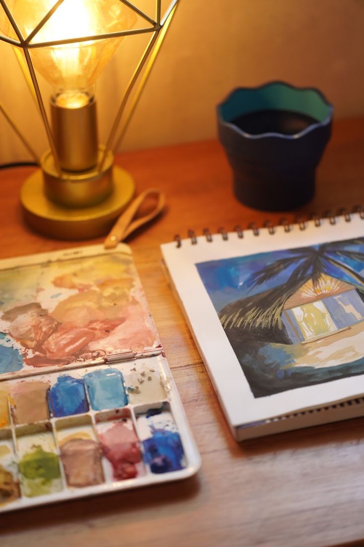Set de acuarelas y una bitácora abierta con un dibujo de una palapa de noche.