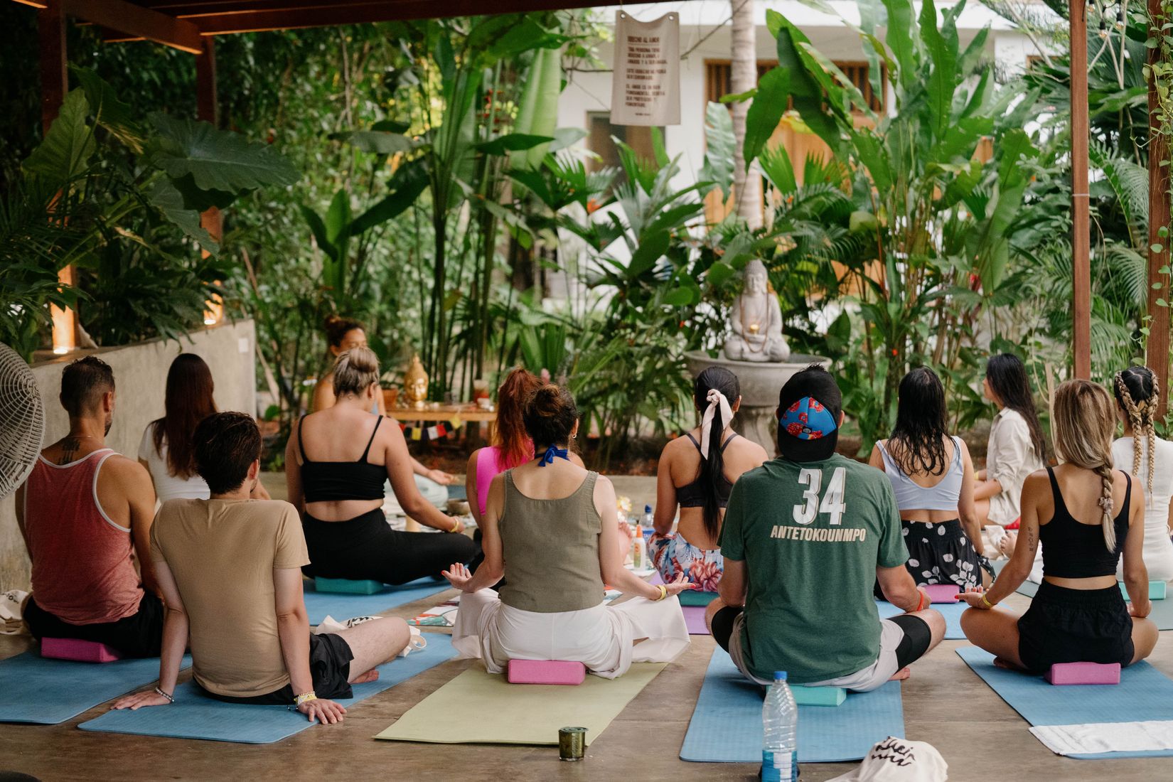 Grupo en clase de yoga sentado en esterillas, entre plantas y una fuente.