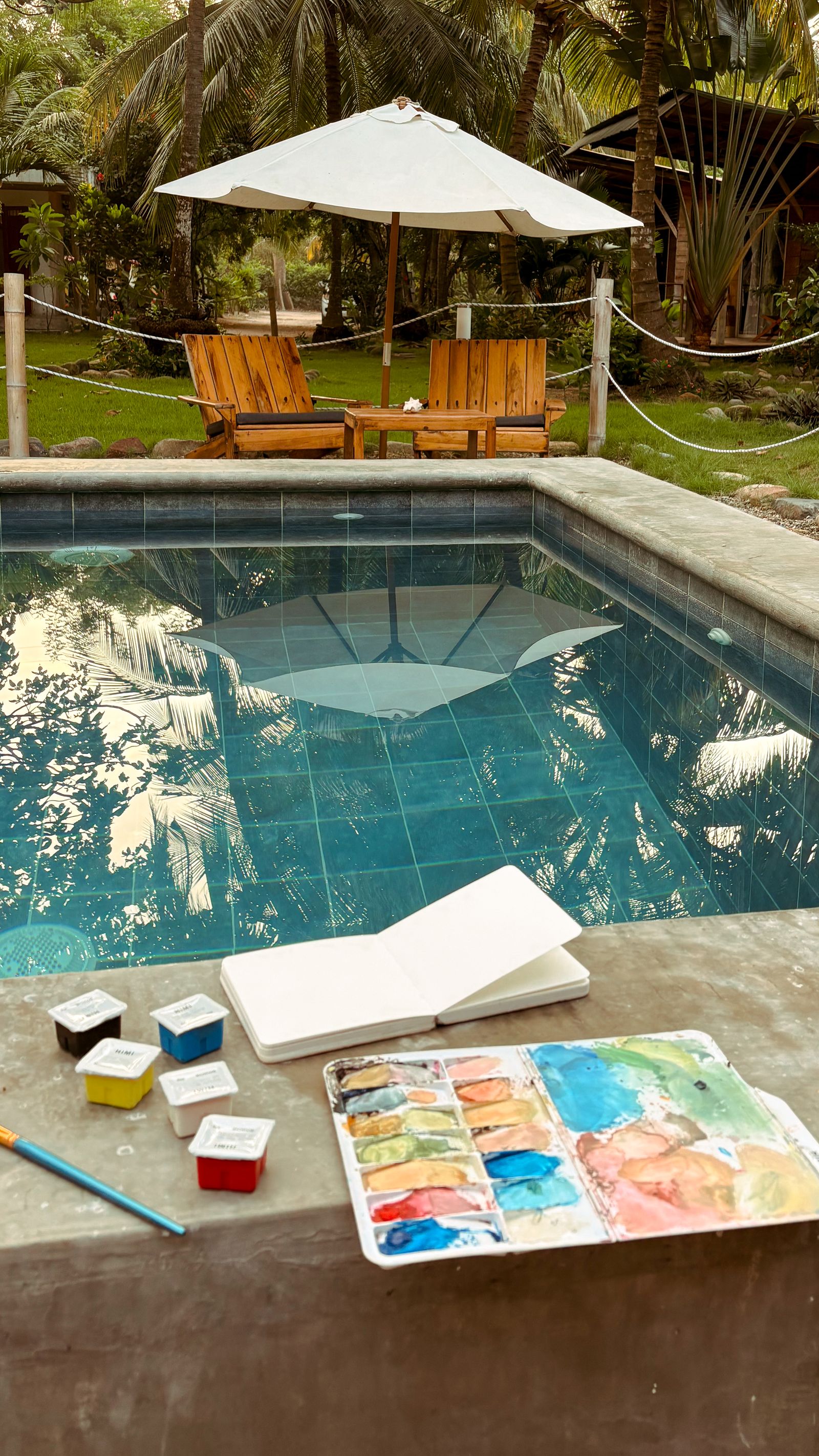 Bitácora, paleta de gouache y materiales de pintura junto a una piscina rodeada de palmeras.