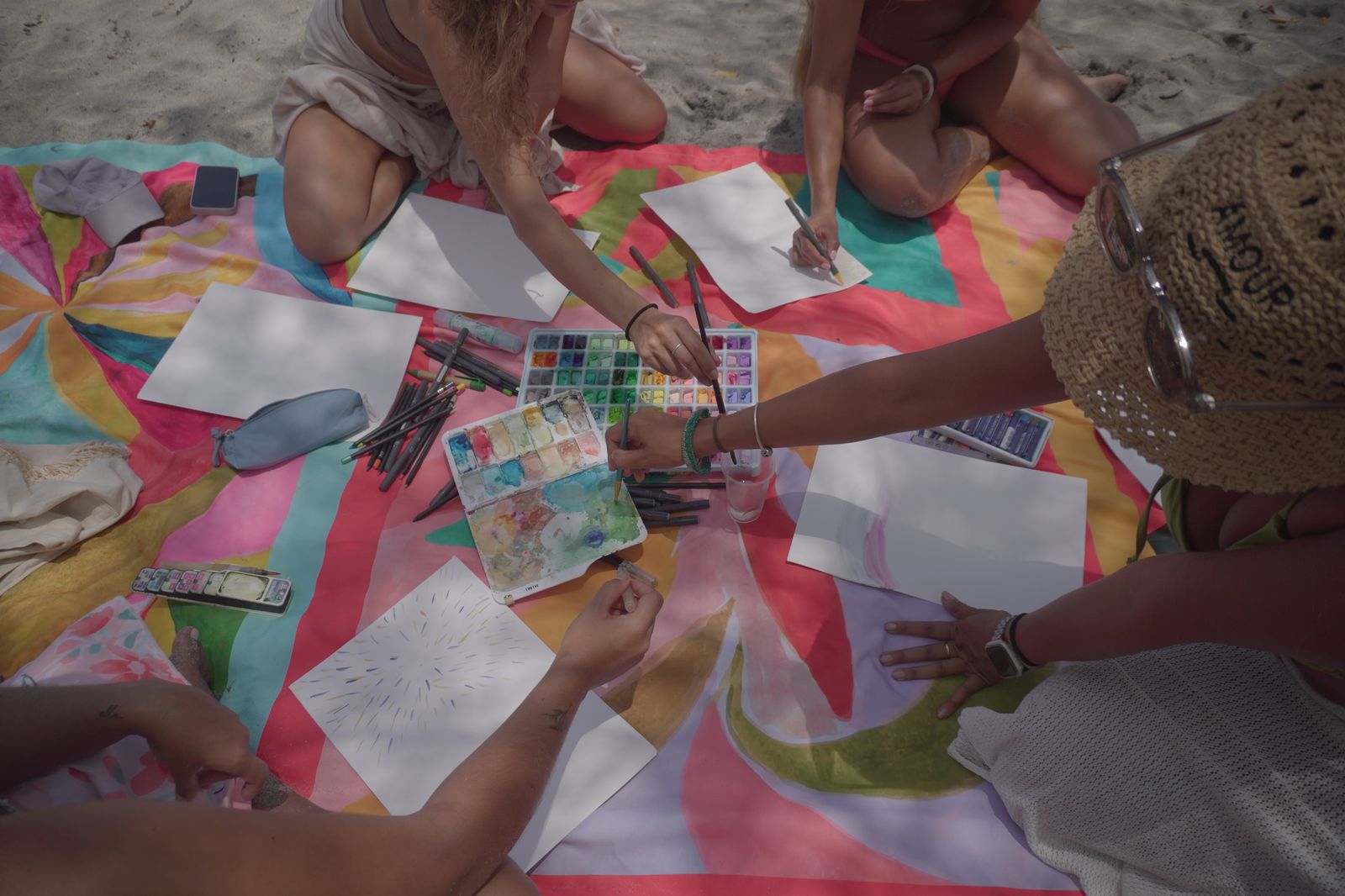 Acuarelas, pinceles y libretas abiertas sobre una manta de colores en la playa.