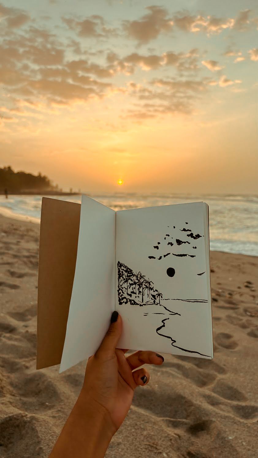 Mano sosteniendo una bitácora abierta con un dibujo de Palomino al atardecer, frente al mar.
